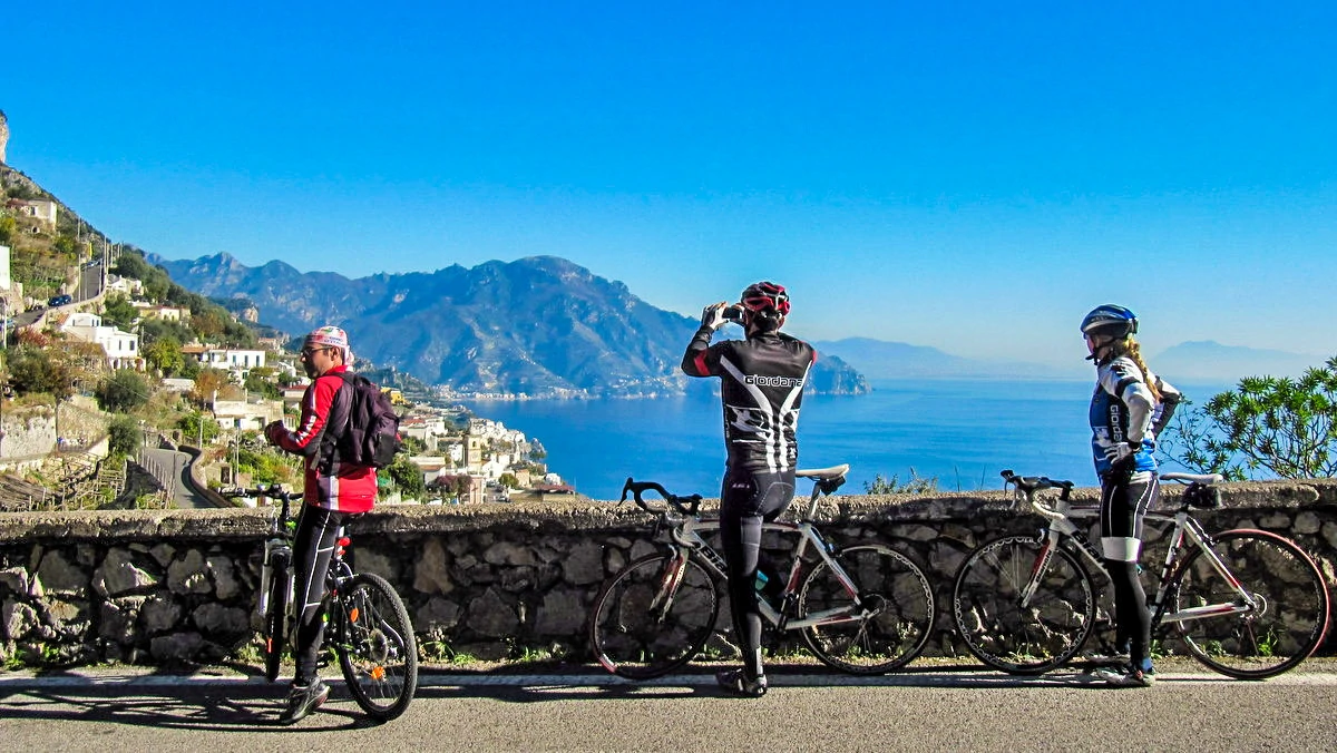 Two cyclists on a scenic road between Positano and Amalfi, overlooking coastal landscape with mountains and sea, under clear blue sky, in cycling gear.