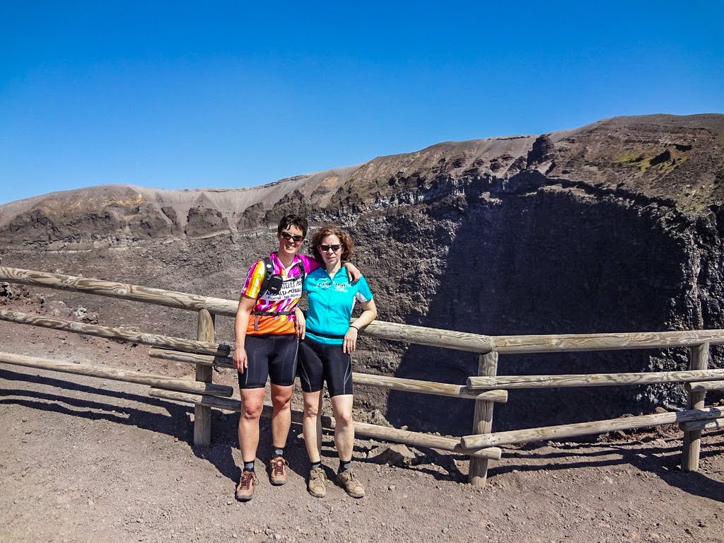 Two people in cycling attire in front of Mount Vesuvius&apos; crater after a guided bike tour, showcasing their adventure with the bike rental service.